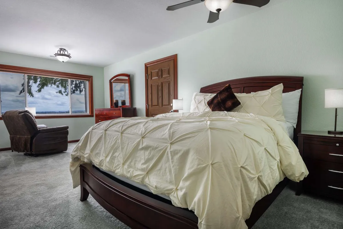 A tranquil bedroom featuring a comfortable bed, an armchair beside a window with panoramic views, and a dresser. The room is bathed in soft natural light, offering a peaceful space for reading or unwinding.