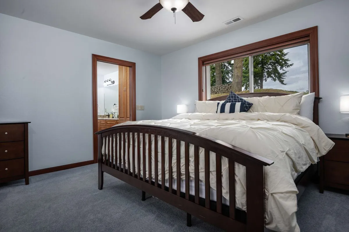 A sunlit bedroom features a comfortable bed, a window framing a gentle outdoor view, a nightstand, and a doorway. The scene exudes relaxation and invites guests to wake up to natural light or enjoy a peaceful night in.