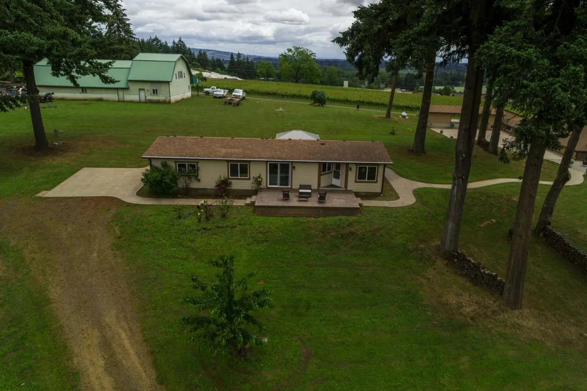 A wide view of the property shows a single-story house with a welcoming wooden deck, surrounded by open lawn, mature trees, and distant rolling hills. The scene captures the feeling of spaciousness and connection to the surrounding landscape, ideal for outdoor play, relaxation, or summer picnics.