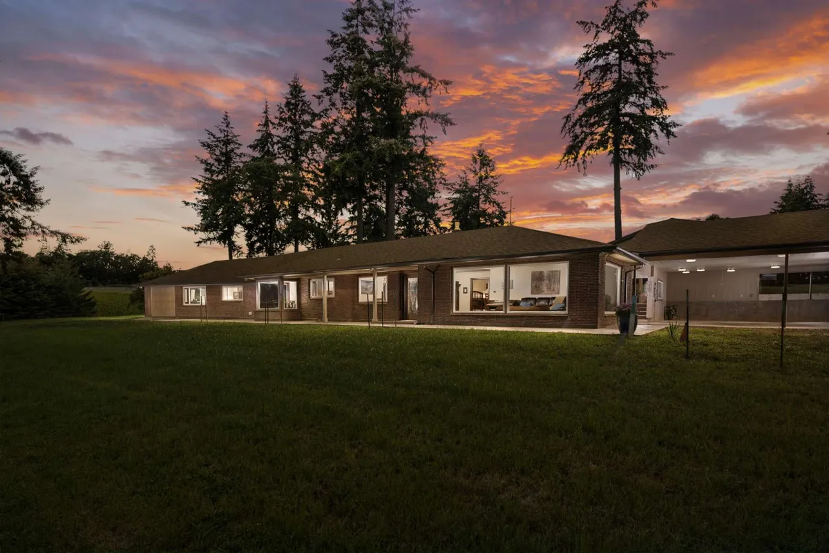 A serene exterior view of the house set against the backdrop of a glowing sunset, with large windows and mature trees framing the scene. The spacious garden area invites outdoor gatherings and relaxation.