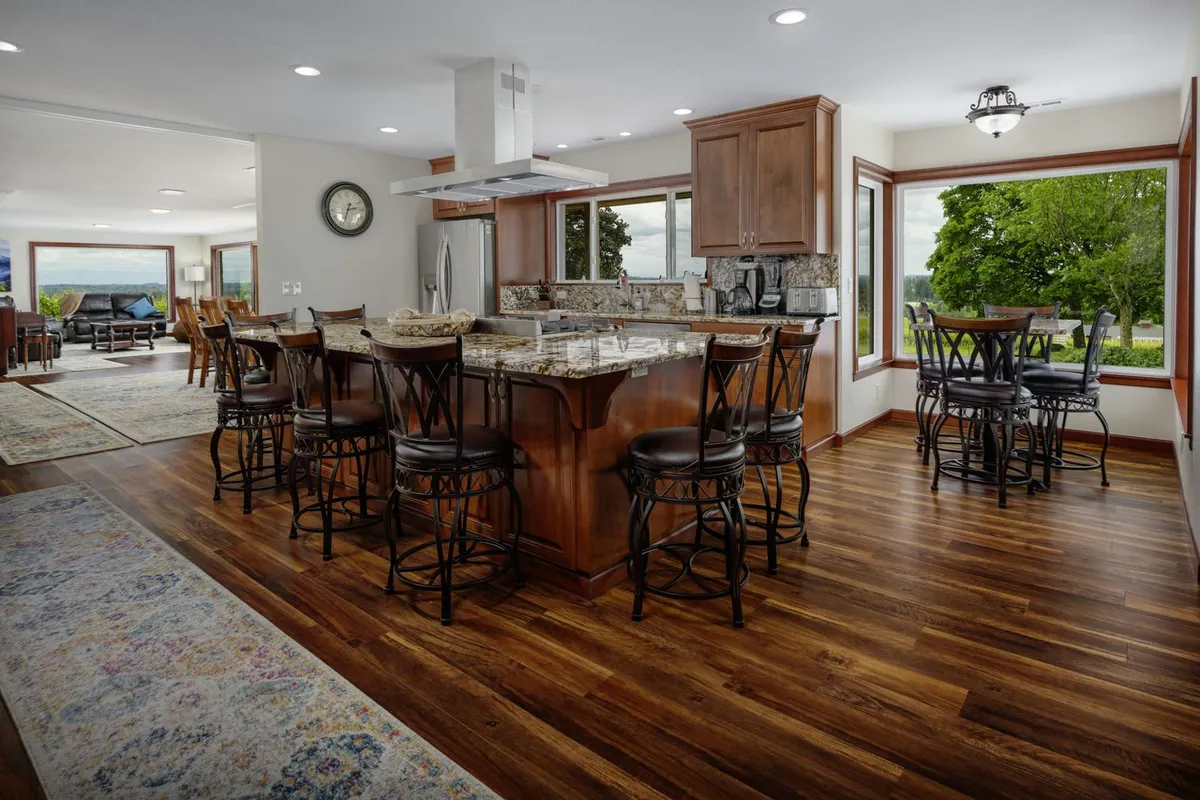 A spacious, modern kitchen with a large granite island, rich wooden cabinetry, and generous windows bringing in natural light. The kitchen seamlessly opens to a cozy dining area with bar stools facing scenic views, evoking a sense of gathering and culinary creativity.