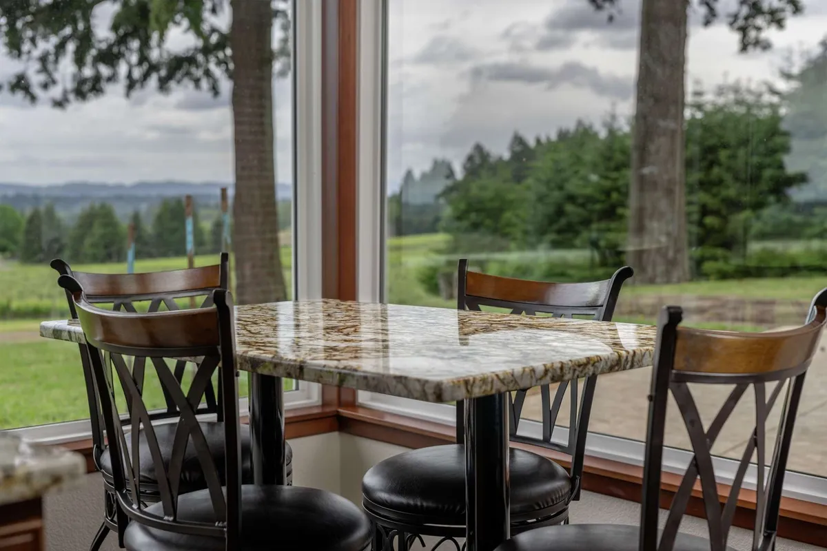 A casual dining area with a granite table and wooden chairs set beside expansive windows that capture the golden hues of sunset over the vineyards. The intimate setting invites social gatherings, coffee mornings, or evenings filled with stories.