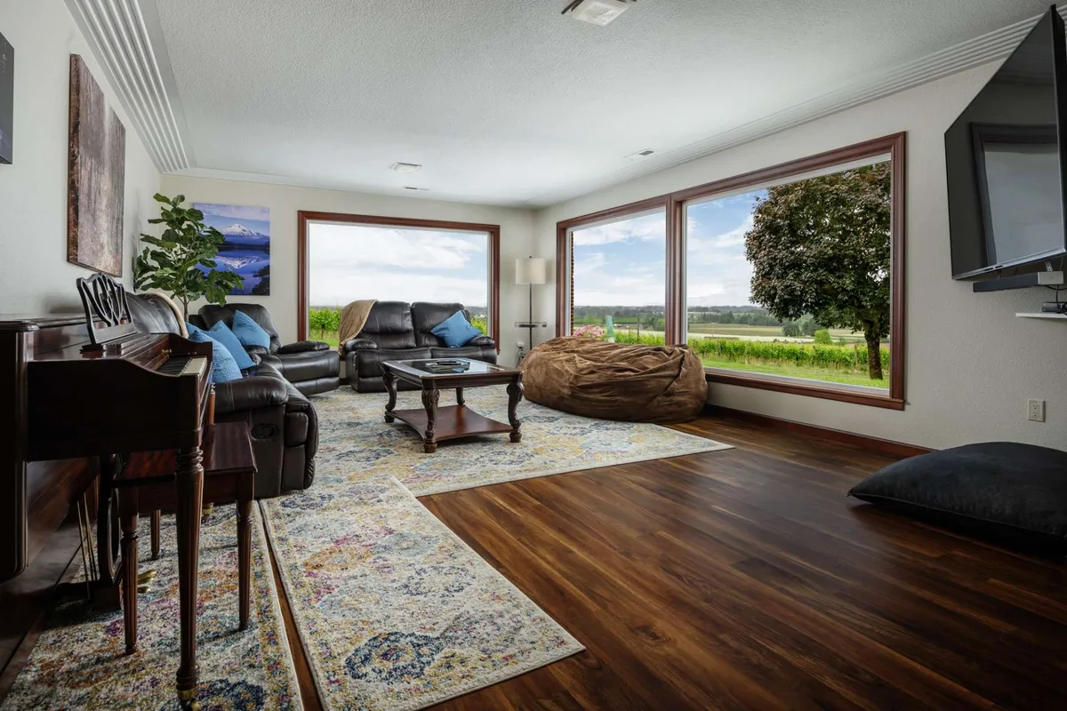 A bright, peaceful living area with oversized, comfortable seating arranged around a decorative rug and a piano. Large windows offer sweeping views of the vineyard and surrounding landscape, filling the space with natural light.