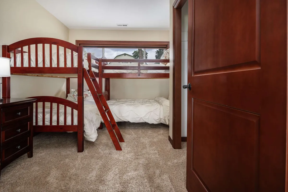 A playful bunk room features twin bunk beds with a cozy nightstand and soft carpet flooring. The ambiance is relaxed and fun, ideal for kids or friends sharing a space and making memories together.