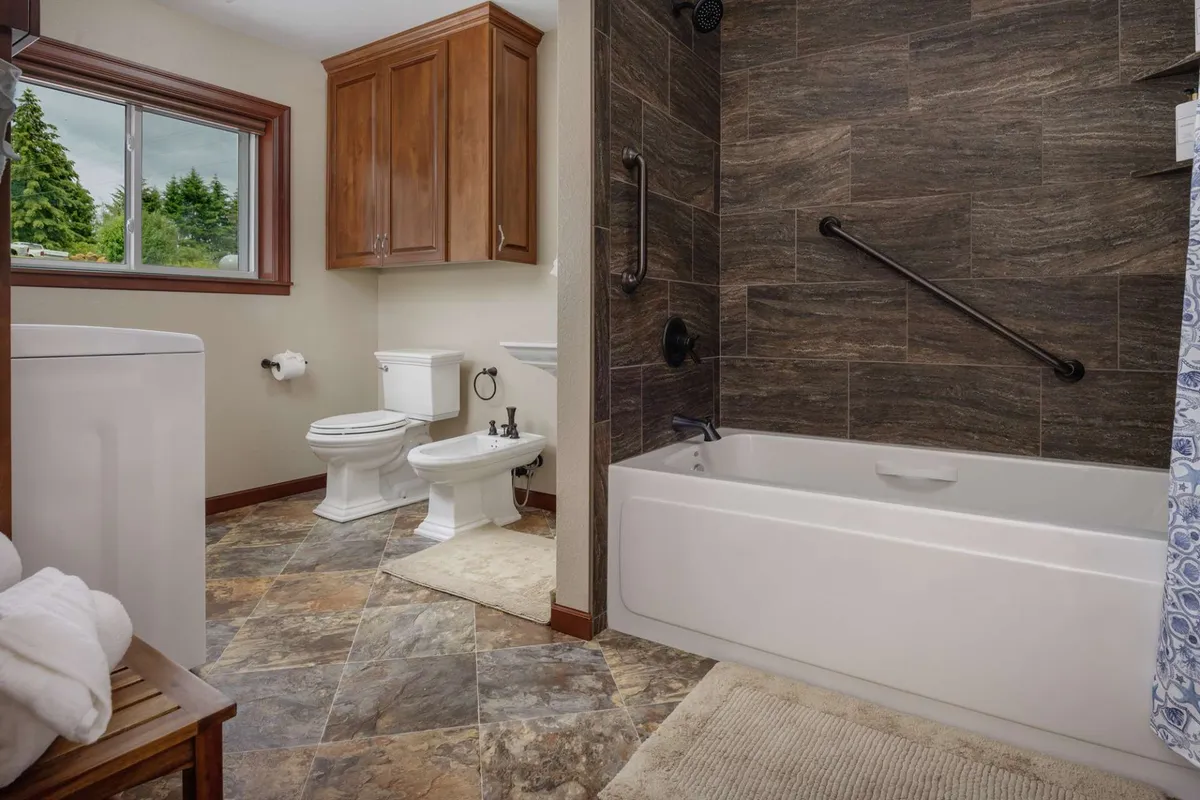 A serene bathroom with a deep soaking tub, separate shower area, and a window offering a peaceful view, complemented by warm wooden cabinetry. The space exudes calm and invites restorative moments of self-care.