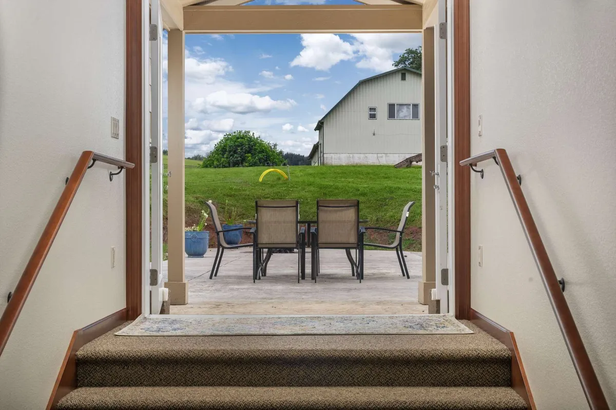 This image features a staircase leading to an outdoor seating area with a green lawn and a barn in the background, seamlessly blending interior access with outdoor living. The inviting patio space, surrounded by natural beauty, suggests opportunities for relaxation, gatherings, and sunset views—key elements for a resort-style escape.