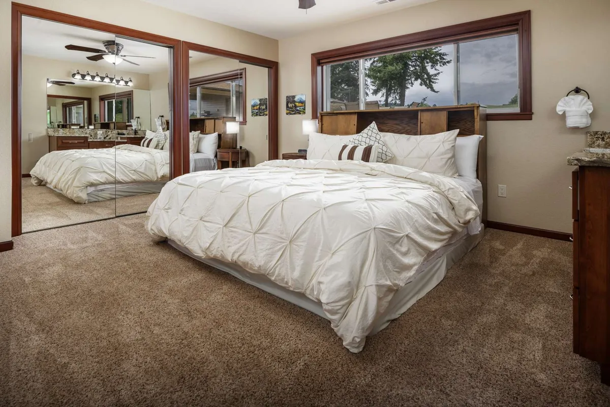 A king-sized bed is dressed with decorative pillows and features a wooden headboard. There is a mirrored closet door and a large window that lets in abundant natural light, offering a tranquil view. The ambiance of the room is peaceful, perfect for restful nights or relaxed mornings.