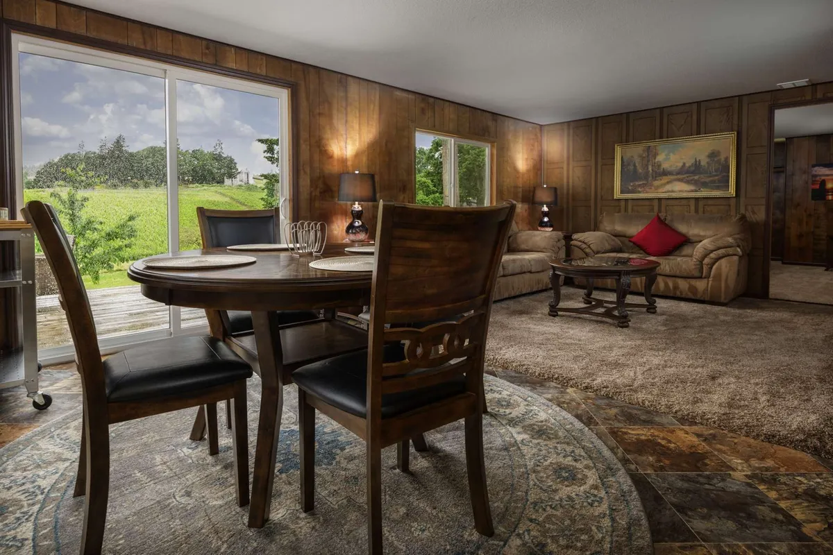A spacious, open-plan living and dining room featuring a large dining table, a comfortable sofa, and expansive windows that frame vineyard views. The scene is decorated with tasteful artwork, creating an inviting and cozy atmosphere ideal for both meals and relaxation.